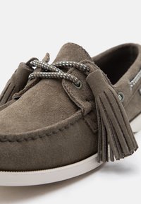 Mocasín de ante marrón con borlas, cordones trenzados y suela de goma blanca, mostrado en primer plano desde un ángulo lateral frontal.