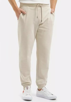 Osoba ubrana w beżowe spodnie dresowe z sznurkiem oraz białe trampki, stojąca z rękami w kieszeniach na jednolitym tle.