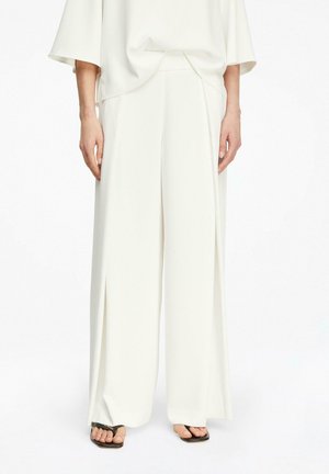 Modelka nosząca luźne białe spodnie o szerokich nogawkach, zestawione z czarnymi sandałami na kwadratowy nosek, stojąca na tle jednolitej białej ściany.