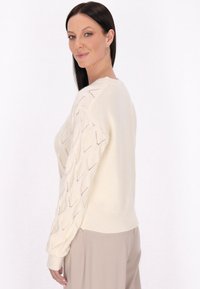 Maglione color crema con un motivo a rombi sulle maniche, polsini a costine e scollatura rotonda. Morbida texture, vestibilità leggermente rilassata.