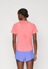 Κοραλλί αθλητικό μπλουζάκι με δίχτυ στο πίσω μέρος, κοντά μανίκια και εφαρμοστή σχεδίαση. Φορεμένο με ανοιχτό μωβ σορτς, παρουσιάζοντας ένα στρωματοποιημένο στυλ.