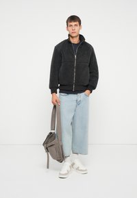 Jachetă neagră din fleece cu fermoar, cu guler înalt, purtată peste o cămașă bleumarin, împreună cu blugi largi din denim deschis și o geantă gri tip crossbody. Tenisi albi.