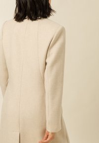 Beige jas met een op maat gemaakte pasvorm, lange mouwen en een gestructureerde stof. Beschikt over naaddetails en een open rugontwerp.