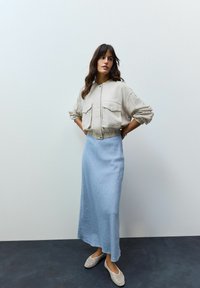 Světlá béžová zkrácená bunda se dvěma kapsami, kombinovaná s modrou maxi suknią a bílými perforovanými slip-on botami. Hladké textury látek.
