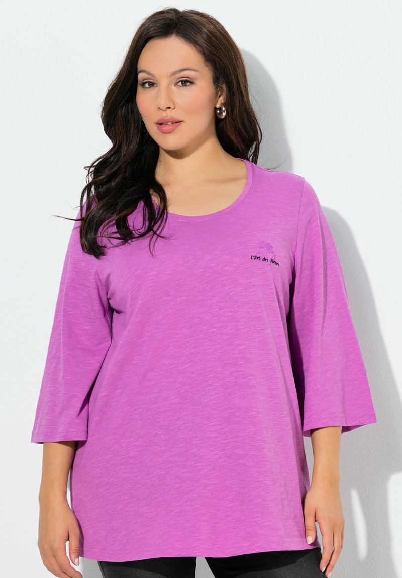 Frau mit langen dunklen Haaren, die ein locker sitzendes lila Oberteil mit dreiviertel Ärmeln trägt, auf dessen Brust kleine Schriftzüge und florale Details zu sehen sind.