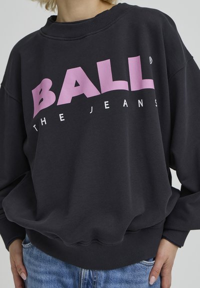 Sweat-shirt noir avec texte graphique rose "BALL" et "THE JEANS", présentant un col rond, une coupe ample et un ourlet côtelé.