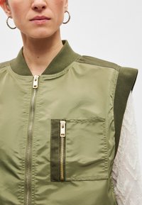 Χακί bomber μπουφάν με φερμουάρ μπροστά και τσέπη, με απαλή λάμψη και ριμπ γιακά, κατασκευασμένο από ελαφρύ, λείο ύφασμα.