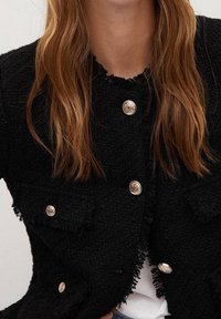 Mustapuku, joka on valmistettu teksturoidusta kankaasta. Siinä on kaksi etutaskua, kultasävyisiä nappeja ja revittyjä reunoja kauluksessa ja helmassa.