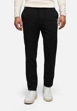Homme portant un pantalon à cordon noir, une chemise blanche à manches longues et des baskets blanches, debout devant un fond uni.