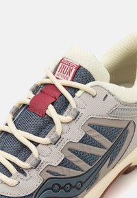 Chaussure de course légère avec un dessus en mesh gris et marine, des accents beiges, des superpositions à motifs et une étiquette rouge avec "saucony RUN anywhere."