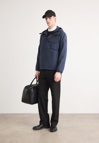 Jachetă bleumarin cu glugă, cu multiple buzunare, purtată peste o cămașă albă cu guler. Pantaloni negri și pantofi, ținând o geantă neagră.