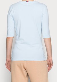 Světle modrý přiléhavý top s třičtvrtinovými rukávy. Hladká textura a zaoblený výstřih, s malým pruhovaným detailem na zadní straně.