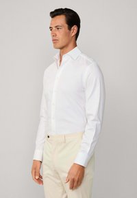 Uomo che indossa una camicia bianca aderente infilata in pantaloni beige, in piedi di lato contro uno sfondo grigio chiaro uniforme.