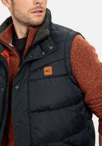 Dunkelblauer Puffer-Weste mit grauem Kragen, ausgestattet mit einem braunen Leder-Logo-Patch und Druckknöpfen; getragen über einem orangefarbenen Strickpullover.