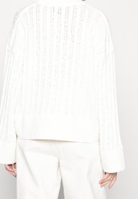 Weißer Strickpullover mit vertikaler Rippenstruktur, locker geschnitten und mit entspannten Ärmeln. Weicher Stoff mit einem samtigen Abschlussband am unteren Saum.