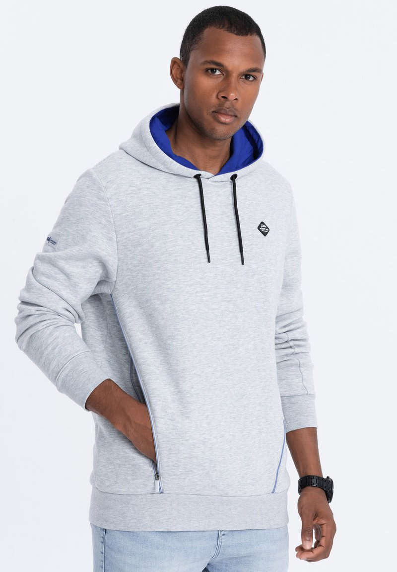 Sudadera gris claro con un forro interior azul, cordones ajustables negros, detalles de cremallera en los laterales y un parche de logo negro en el pecho.