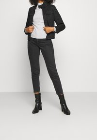 Veste en denim noir avec deux poches poitrine, pull gris en dessous, jean skinny noir et bottes noires à talons. Design épuré et ajusté.
