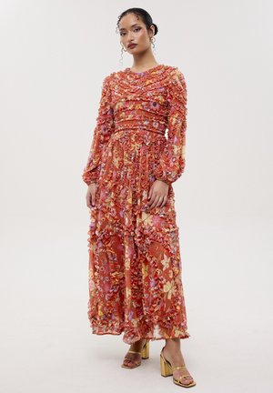 Mujer con vestido rojo de flores de manga larga y largo hasta el tobillo con volantes y sandalias doradas de tacón, de pie contra un fondo liso y claro.