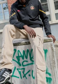 Svart zip-up jacka med Atlanta United-logotyp, beiga cargobyxor med dragkedjefickor, grå sneakers med vita ränder, sitter på en strukturerad yta.