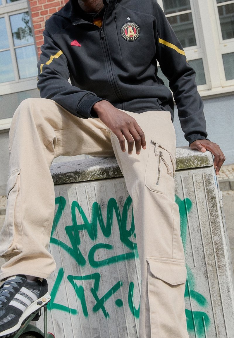 Svart zip-up jacka med Atlanta United-logotyp, beiga cargobyxor med dragkedjefickor, grå sneakers med vita ränder, sitter på en strukturerad yta.