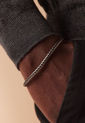 Geweven zilveren armband om een donkere huidpols, met een textuur van een zwarte trui manchette zichtbaar erboven. Glad metaal met gevlochten ontwerpelementen.