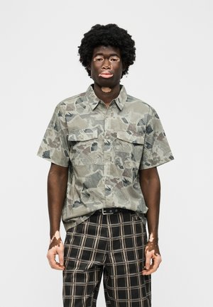 Muž s vitiligem, který má na sobě krátký rukáv zelené maskáčové tričko a černé kostkované kalhoty, na bílém pozadí.