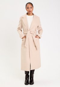 Beige wool blend long coat with a belted waist, notched lapels, and two front pockets; paired with black knee-high boots.