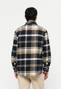 Camicia di flanella a quadri neri, bianchi, grigi e beige. Presenta maniche lunghe, polsini con bottone e orlo arrotondato. Tessuto texturizzato con una finitura morbida.
