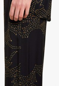 Tessuto nero con motivo di cuciture dorate, caratterizzato da forme astratte e linee curve, con un primo piano sulla mano che tiene il capo.