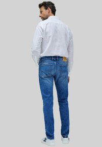 Vaqueros de mezclilla azul descoloridos con corte recto, con bolsillos traseros y una etiqueta de cuero marrón en la cinturilla. Camisa blanca de botones puesta.