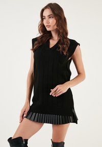 Black knit sweater dress featuring a V-neck, ribbed texture, and short sleeves, with a pleated faux leather skirt at the hem.