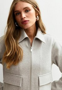 Lichtgrijze zip-up jas van zacht materiaal, met een kraag, voorzakken en een gladde textuur.