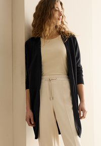 Svart cardigan med långa ärmar över en ljusbeige topp. Outfiten har en avslappnad design och kompletterande beige byxor med elastiskt midjeband.