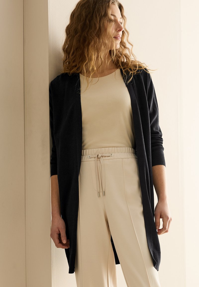 Svart cardigan med långa ärmar över en ljusbeige topp. Outfiten har en avslappnad design och kompletterande beige byxor med elastiskt midjeband.