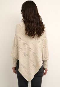 Beige poncho met een gestructureerd breipatroon en zigzagdetail. Heeft een asymmetrische zoom en wijde mouwen, gedragen over donkere jeans.
