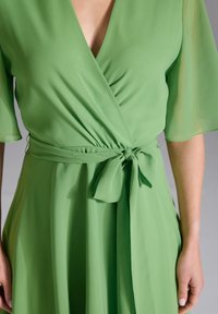 Robe portefeuille verte en tissu léger, dotée de manches larges, d'une ceinture nouée à la taille et d'un décolleté en V profond. Texture lisse, design fluide.