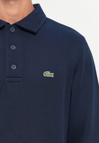 Navyblaues Langarm-Polohemd mit drei Knöpfen und grünem Krokodil-Logo auf der linken Brust.
