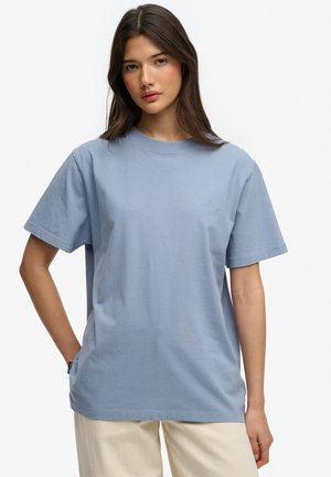 Ung kvinde med langt brunt hår iført en lyseblå løs T-shirt og cremefarvede bukser, stående med den ene hånd i lommen.