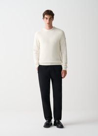 Suéter de punto crema con cuello y puños acanalados, combinado con pantalones negros. Modelo de pie sobre un fondo liso, con zapatos negros.