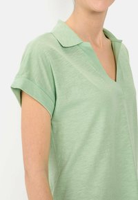 Lichtgroene polo met korte mouwen en een v-hals ontwerp. Zachte textuur, opgerolde cuffs en een subtiel gemêleerd patroon.