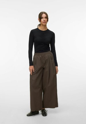 Žena s hnědými vlasy staženými do culíku, oblečená v černém nariaseném tričku s dlouhým rukávem, volných hnědých kalhotách s širokými nohavicemi a černých šněrovacích botách na bílém pozadí.