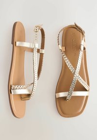 Gouden platte sandalen hebben een gevlochten strapdesign met gladde metallic accenten en een bruine textuurloze voetbed; open teen met een minimalistische vorm.