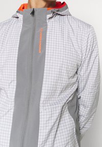 Veste légère à capuche blanche avec motif à carreaux gris. Comprend un panneau avant gris et des accents orange vif. Fermeture éclair et capuche ajustable.