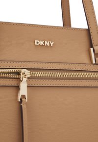 DKNY béžová taška s textúrovaným materiálom, zlatým zipsom a logom. Obsahuje precízne švy a štruktúrovaný dizajn.