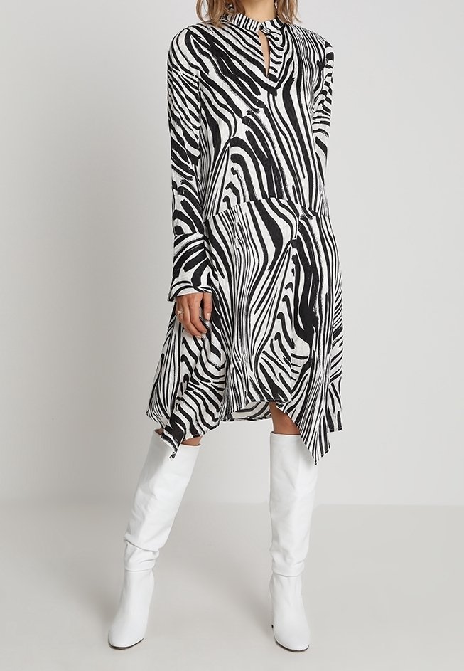 Robe noire et blanche à rayures zèbres avec un décolleté en trou de serrure, manches longues, ourlet asymétrique, portée avec des bottes blanches hautes jusqu'aux genoux.