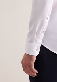 Biała koszula z teksturowanej tkaniny, z ostrym mankietem i perłowymi guzikami. Rękaw zdobi haftowane logo.