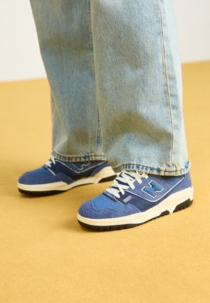 Sneaker blu e bianchi con una tomaia testurizzata e multitonale, caratterizzate da un logo prominente e una suola in gomma nera, abbinate a jeans chiari.