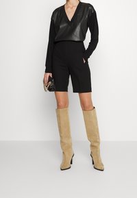 Svart långärmad topp med V-ringning, kombinerad med skräddarsydda svarta shorts. Beigea knähöga stövlar med klack, svart clutch med guldkedjedetalj.