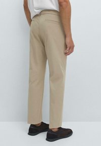 Homme portant un pantalon beige droit et des sneakers marron foncé, se tenant devant un fond gris clair uni.