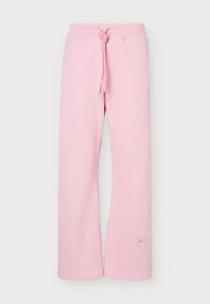 Femme debout, de face, portant un t-shirt court blanc, un pantalon de survêtement rose à jambes larges et des baskets blanches sur un fond gris uni.